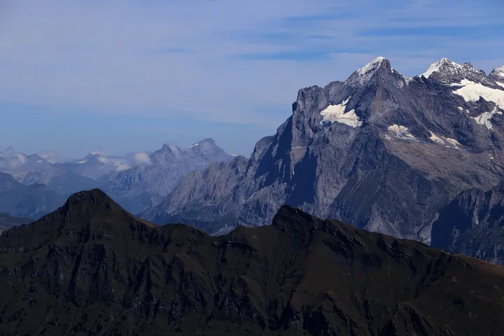 Snow-capped Swiss Alps rising above dark foreground ridges beneath a blue sky.