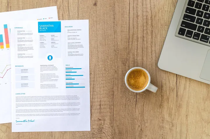 Wydrukowane CV na drewnianym biurku obok wykresów, rogu srebrnego laptopa i białej filiżanki espresso