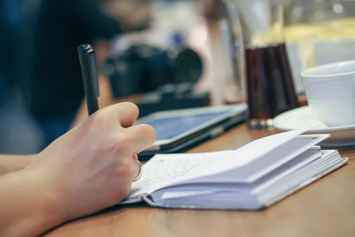 Dłoń z długopisem pisząca w notesie na drewnianym biurku obok filiżanki kawy