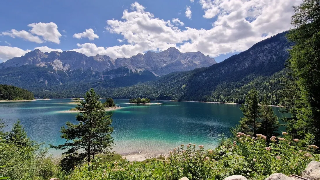 Turquoise mountain lake with forested shores, alpine peaks, and wildflowers beneath a cloudy blue sky.