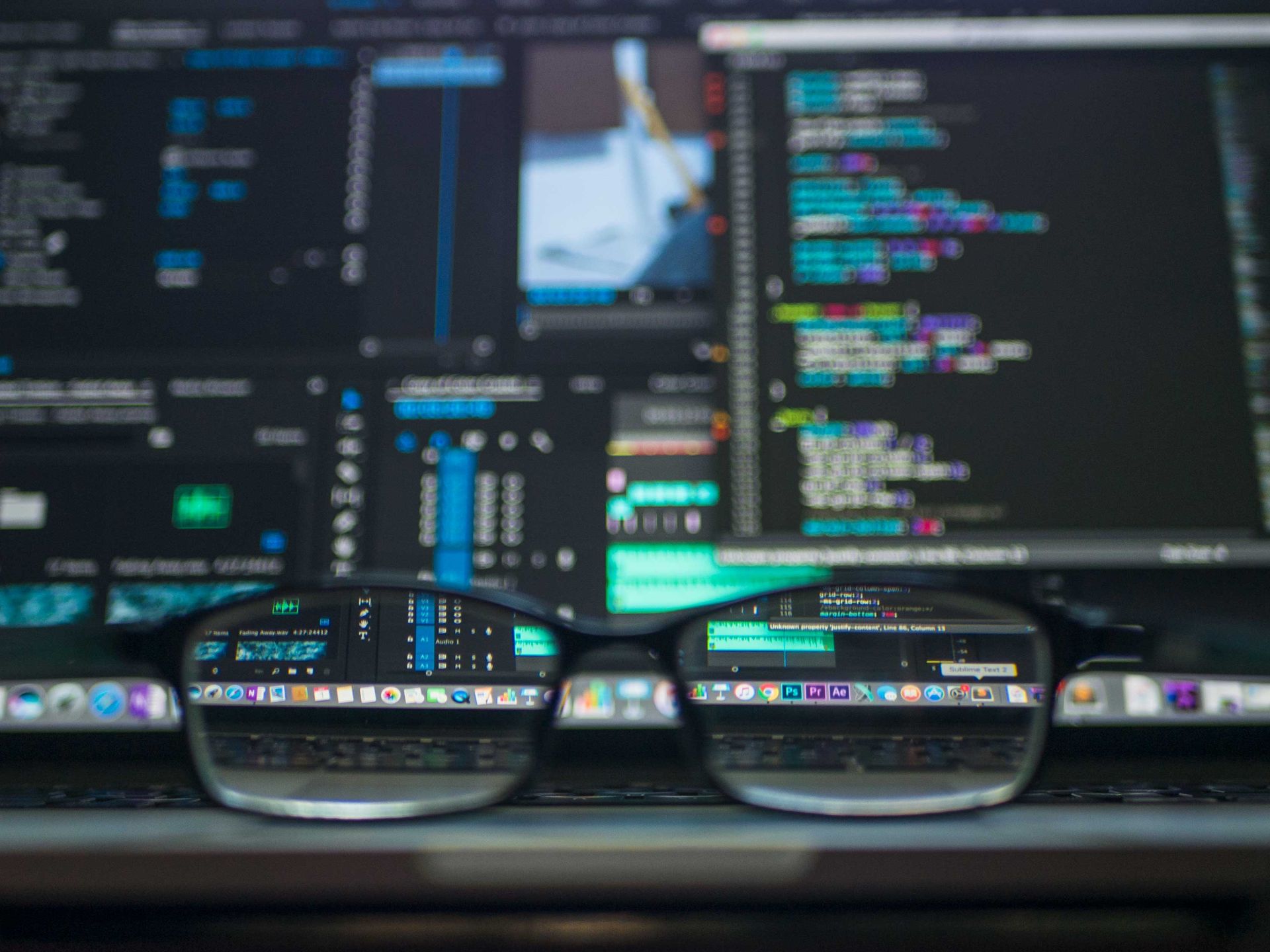 Black-framed glasses resting on a laptop keyboard with code reflected in the lenses