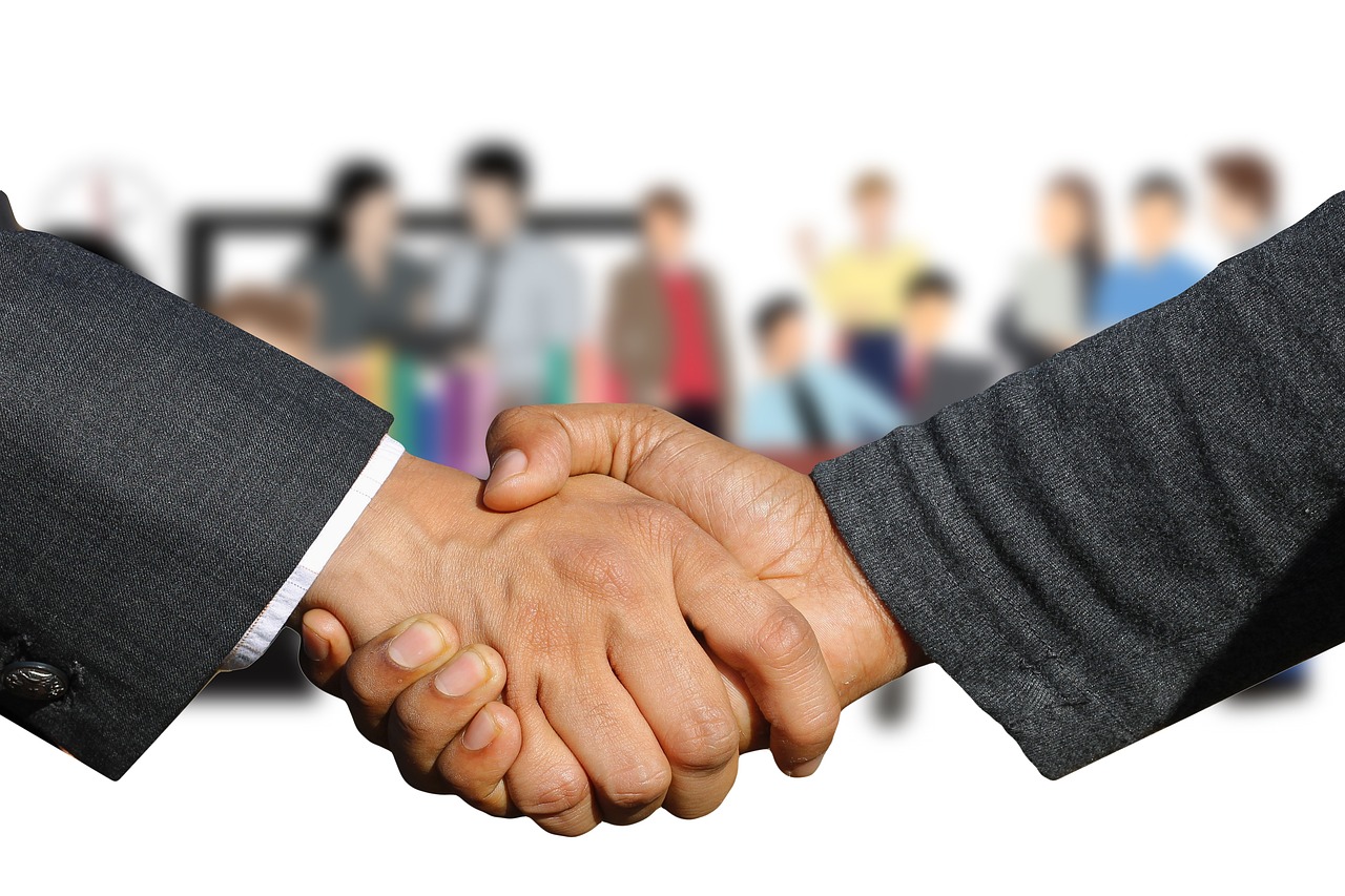 Close-up of two people in dark suit jackets shaking hands, with a blurred office meeting in the background