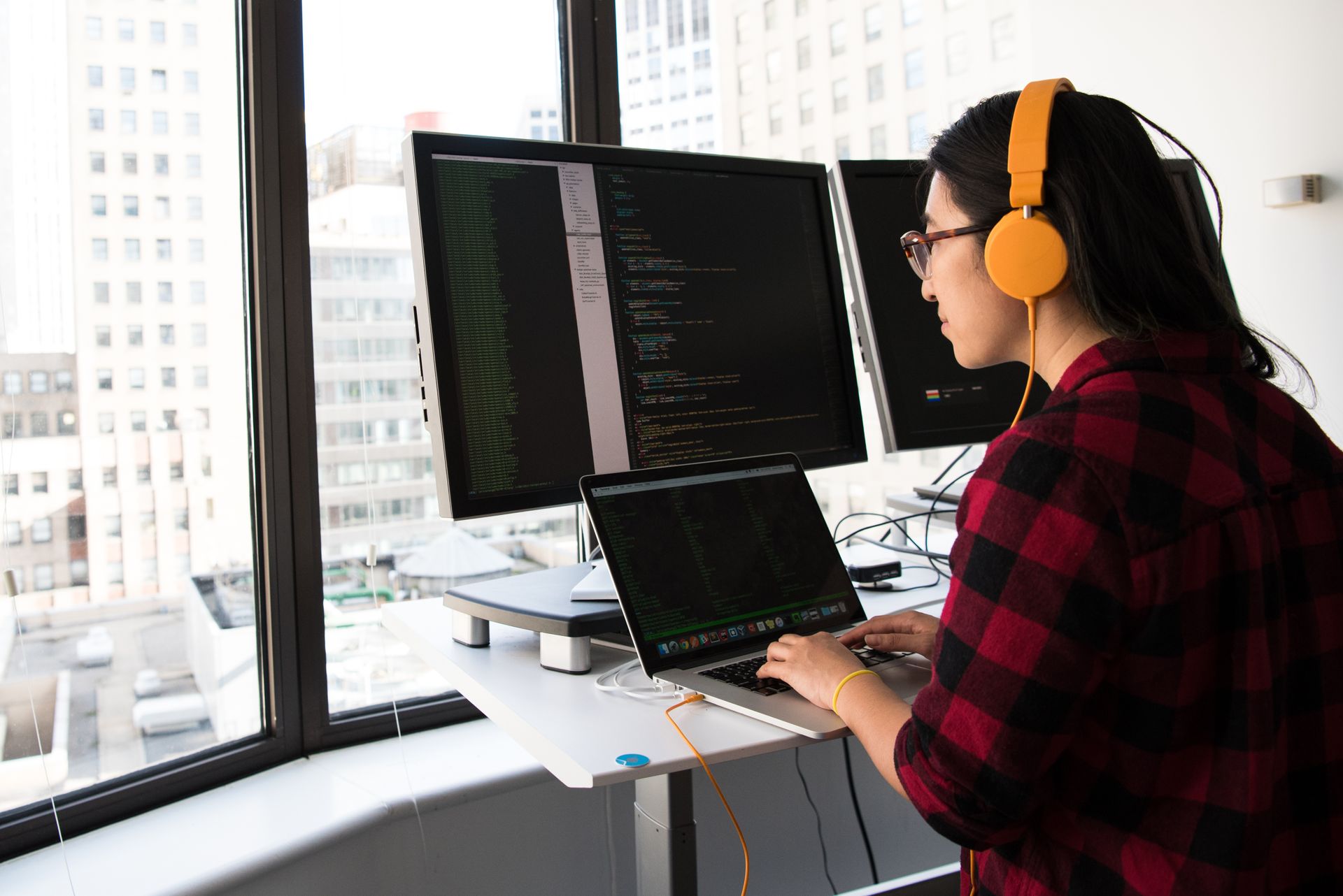 Developer wearing headphones typing on a laptop beside two monitors displaying code