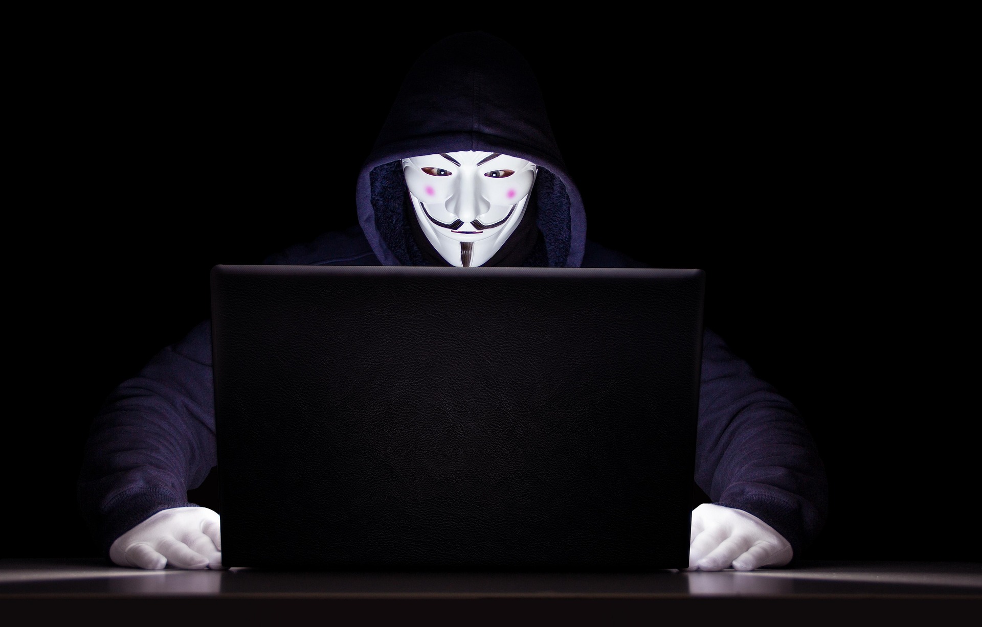 A hooded figure wearing an anonymous mask sitting behind a laptop in a dark setting