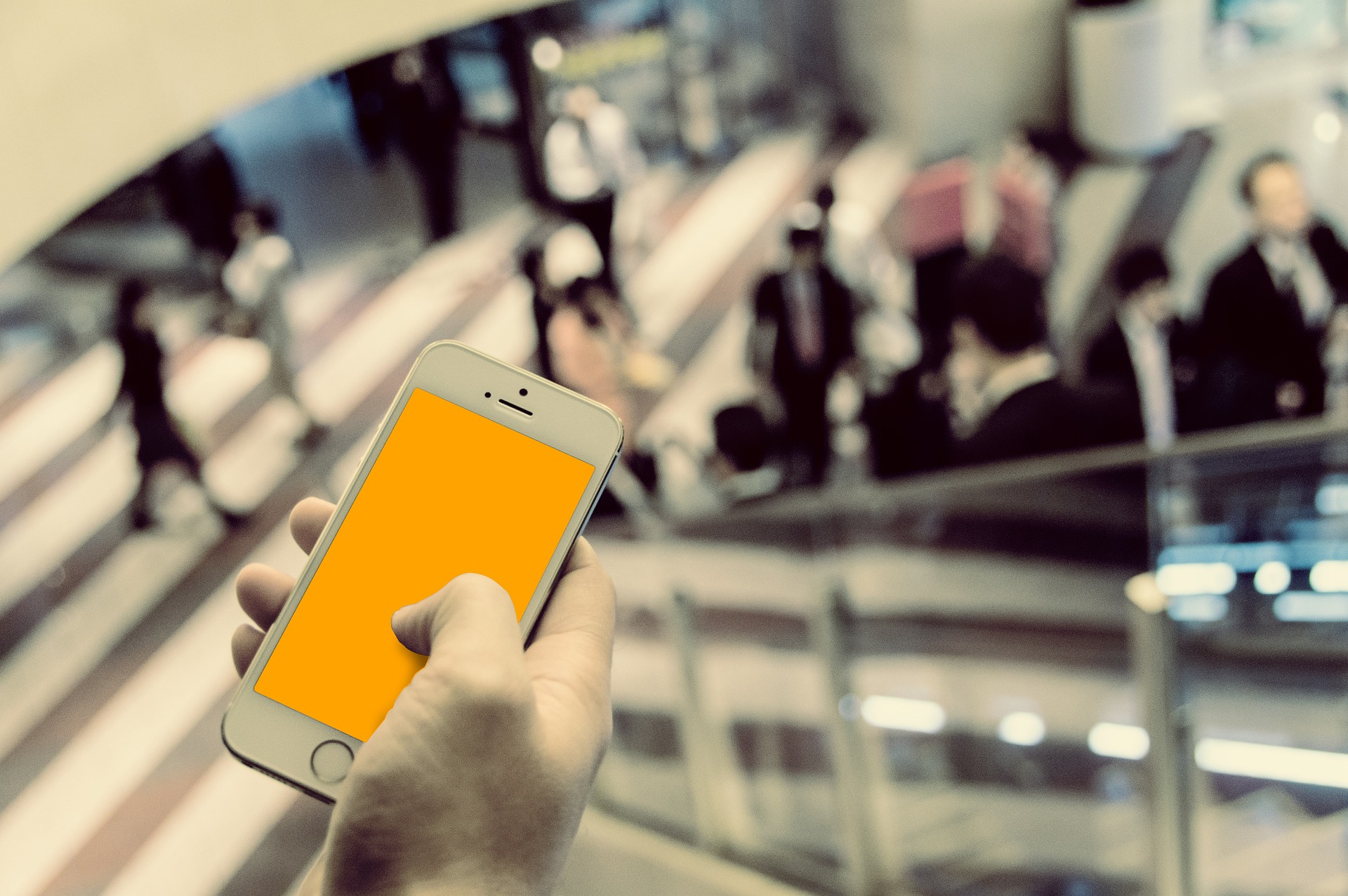A hand holding a smartphone with an orange screen above a blurred busy public space with people on escalators