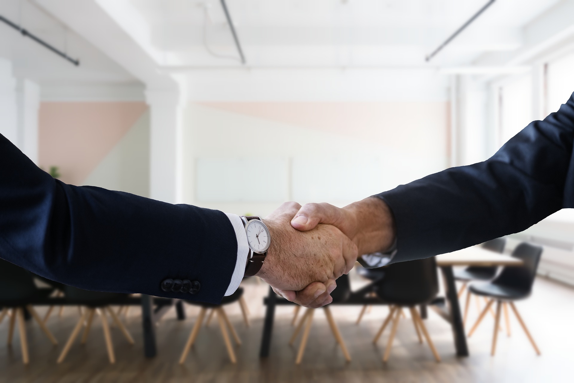 Two people in dark suits shaking hands in a bright modern office with chairs and a table in the background