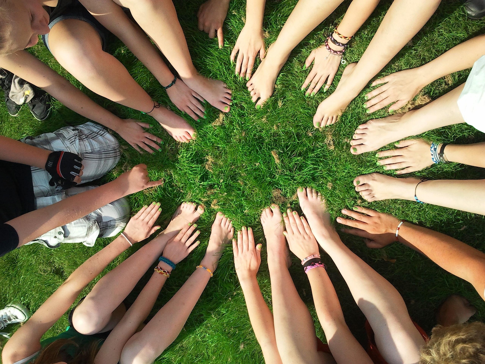 A group of people sitting in a circle on grass with their hands and bare feet reaching toward the center