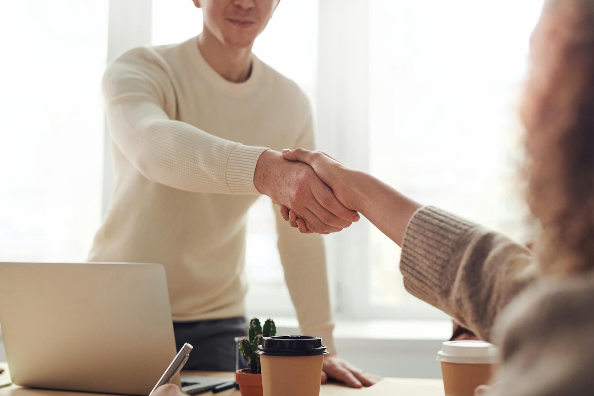 Two people shaking hands over a wooden desk with a laptop, a small cactus, and takeaway coffee cups