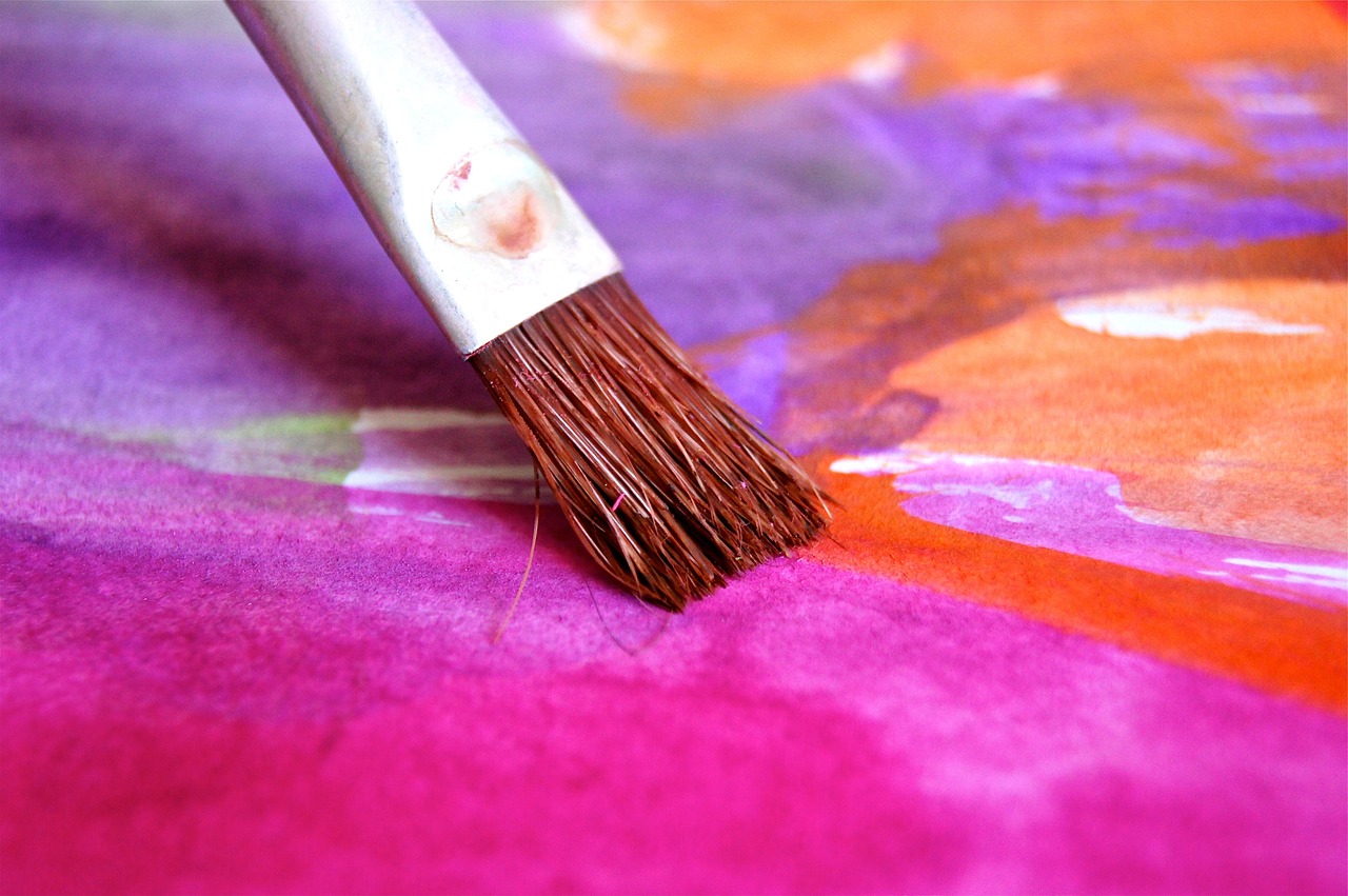 Close-up of a paintbrush touching wet watercolor paint in pink, orange, and purple on paper