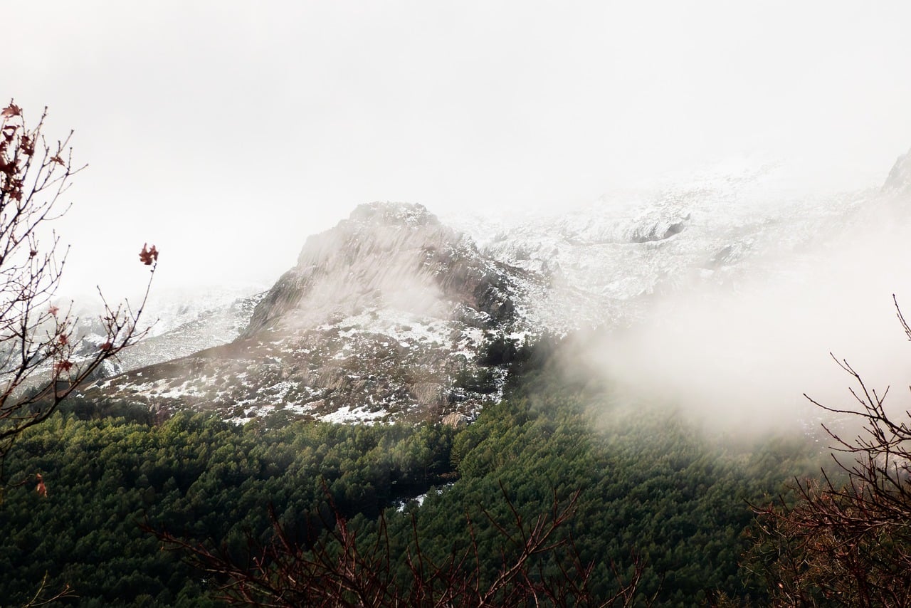 A snow covered mountain with fog drifting across the forest below it