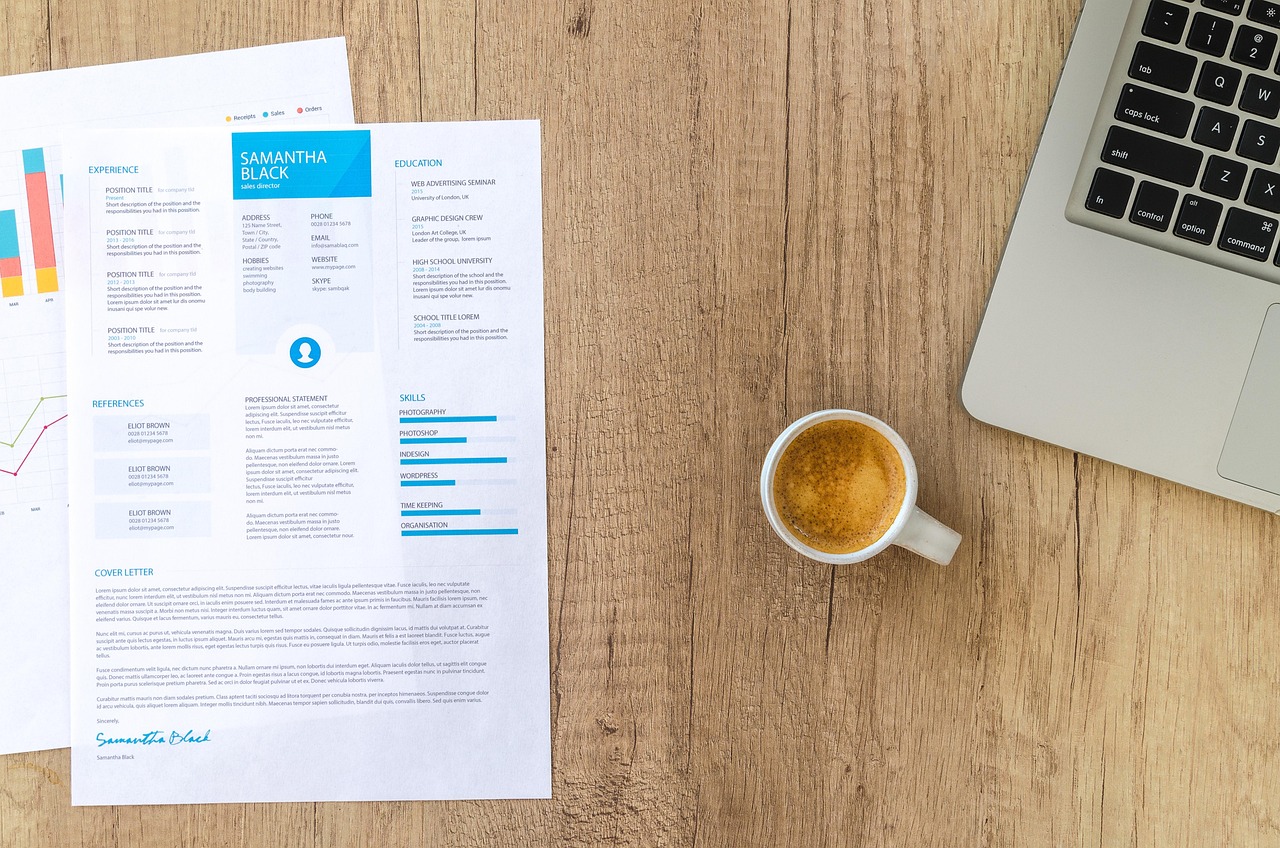 Printed resume on a wooden desk beside charts, a silver laptop corner, and a white cup of espresso