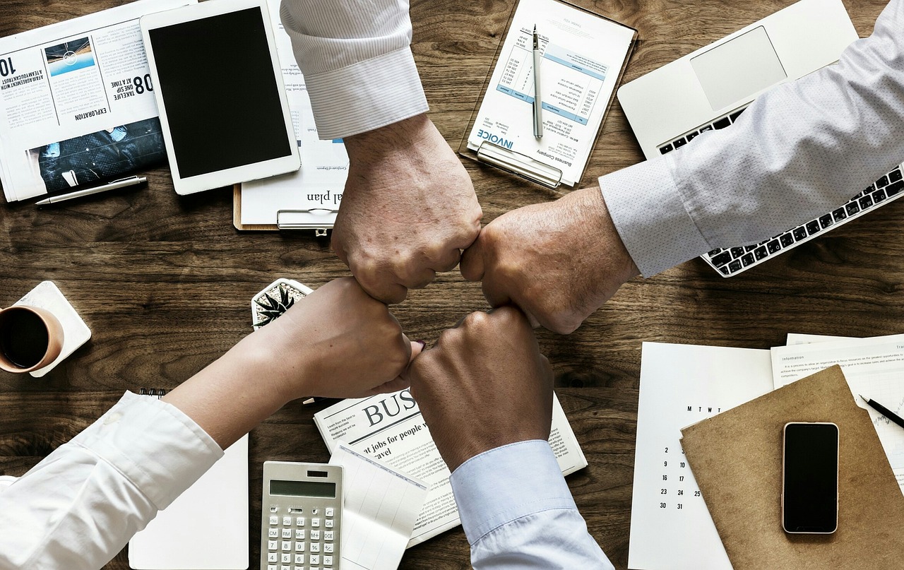 Four hands doing a fist bump over a wooden desk surrounded by a tablet, laptop keyboard, smartphone, calendar, and documents