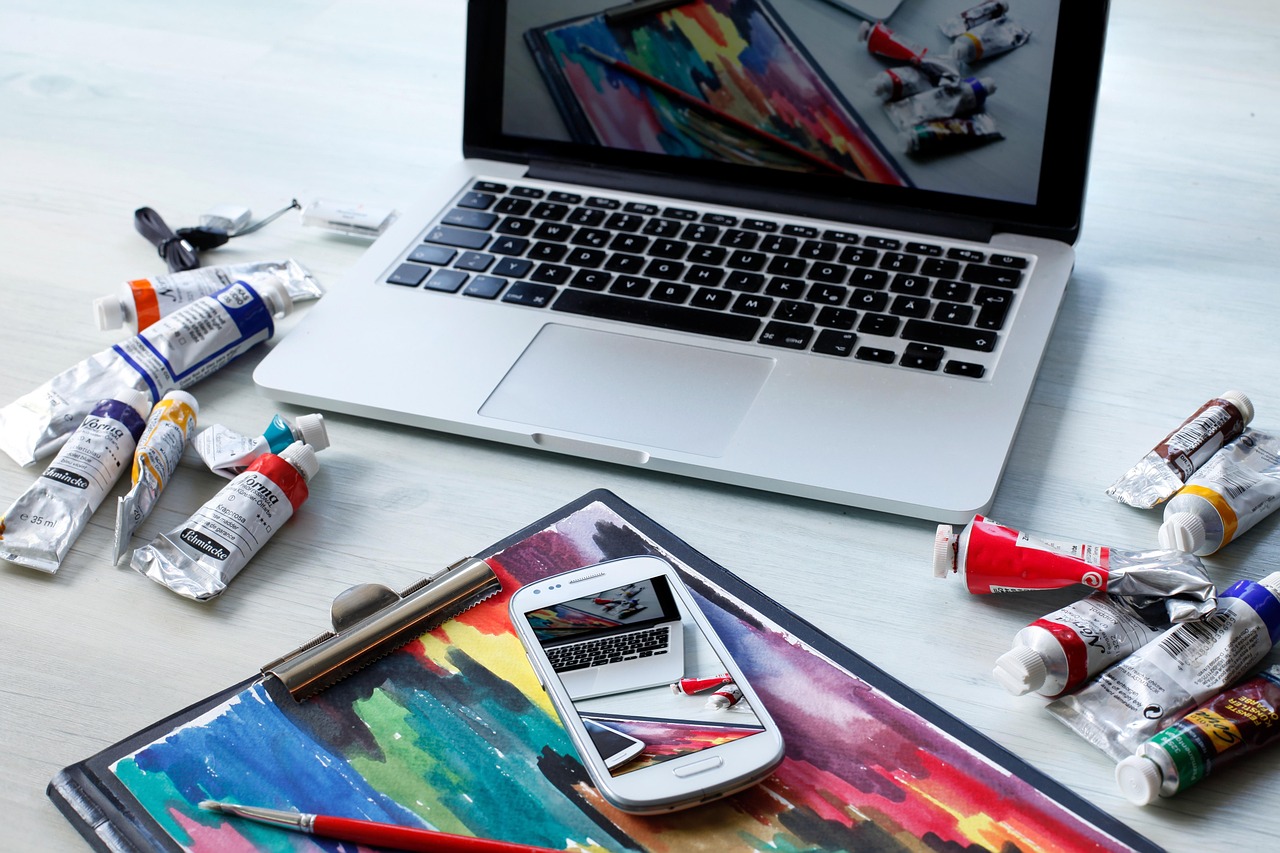 An open laptop surrounded by paint tubes and a watercolor sketchbook, a colorful digital painting visible on screen