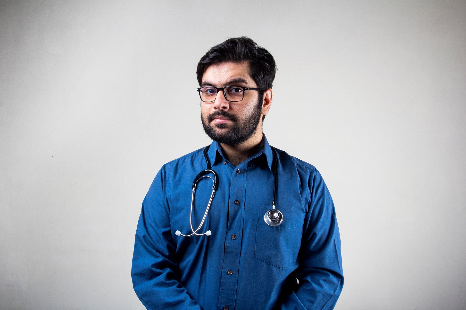Man in a blue shirt and glasses with a stethoscope around his neck posing against a plain background